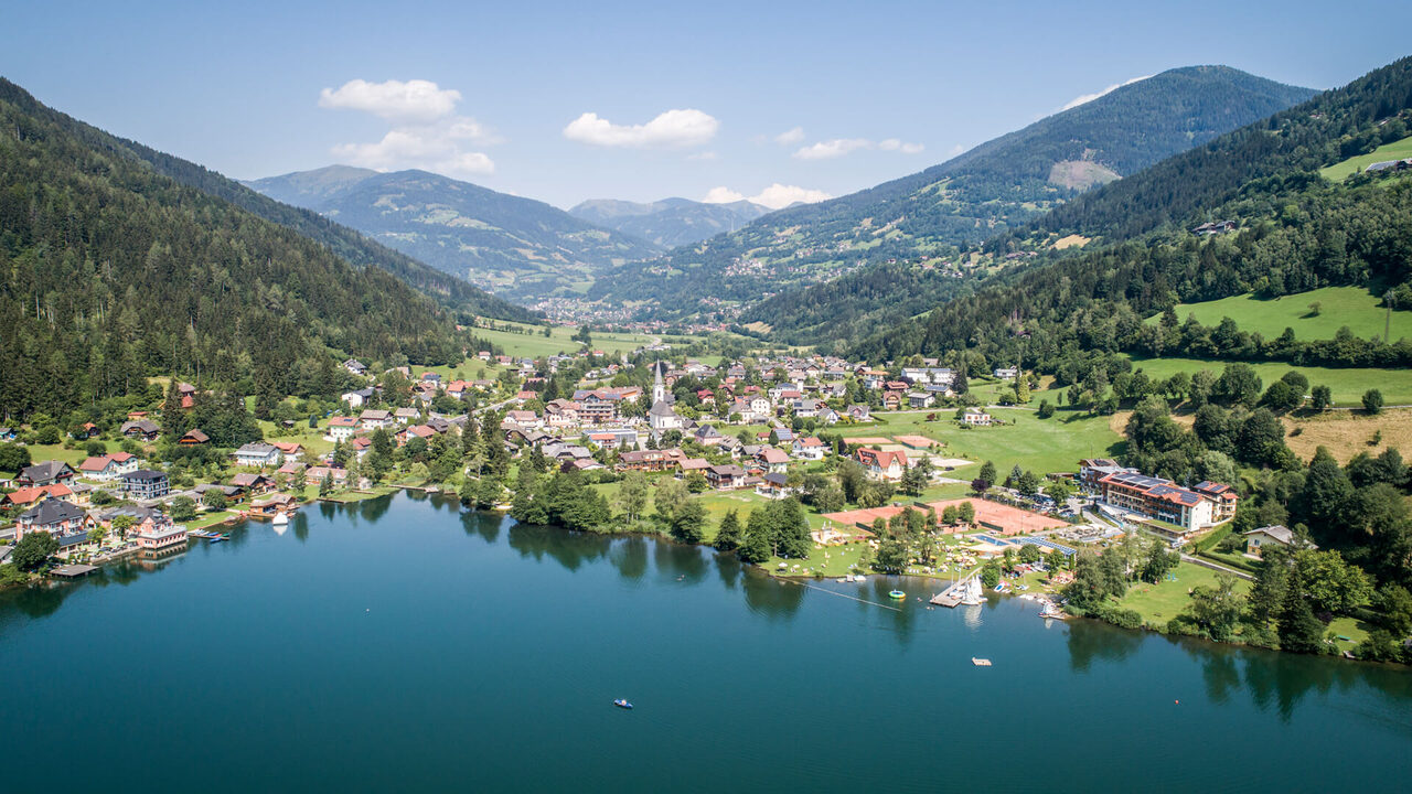 Luftaufnahme eines Ortes am See, umgeben von bewaldeten Bergen